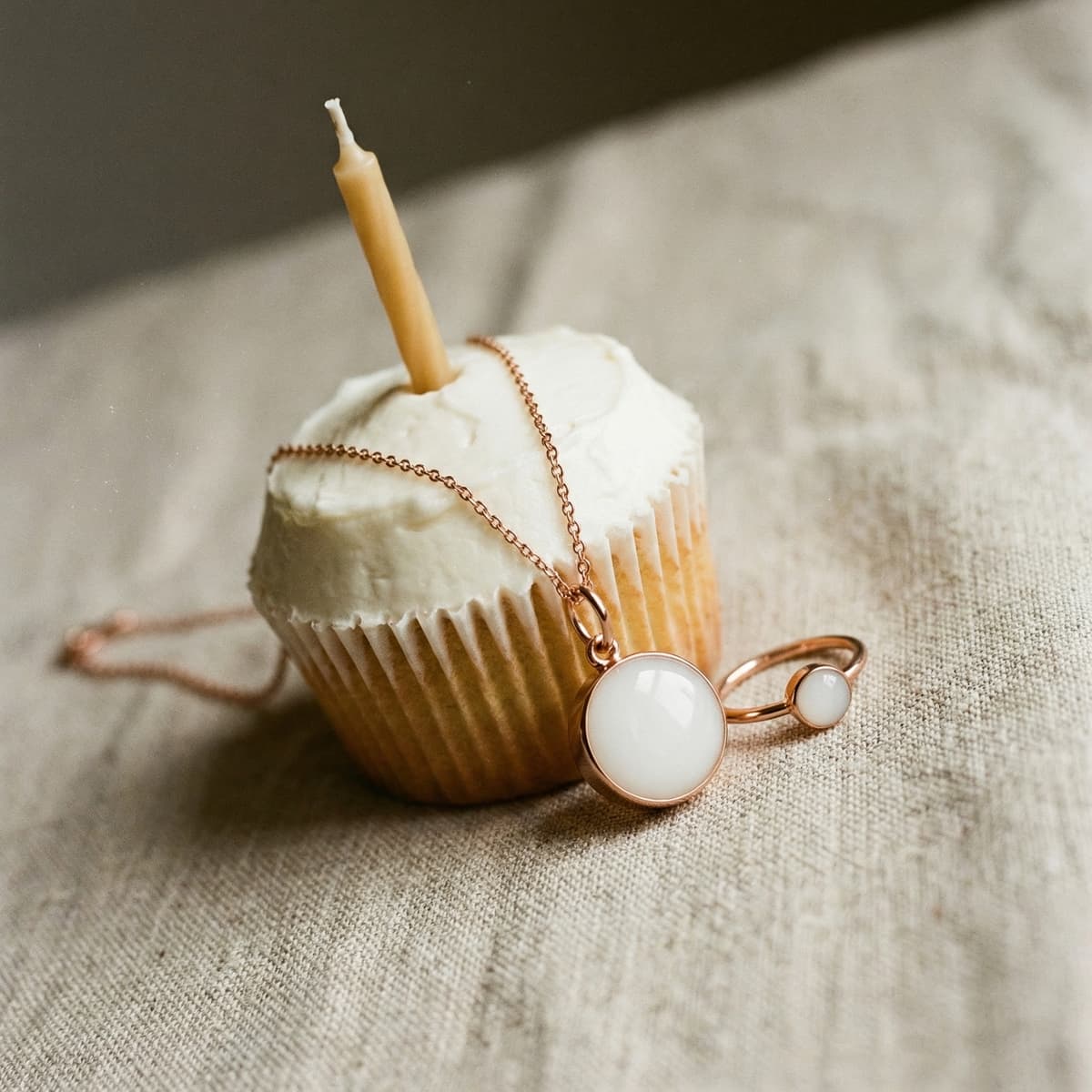 Vanilla cupcake with a single candle beside rose-gold breastmilk jewelry pendant and ring with milky white stones, celebrating a 1 year breastfeeding award keepsake