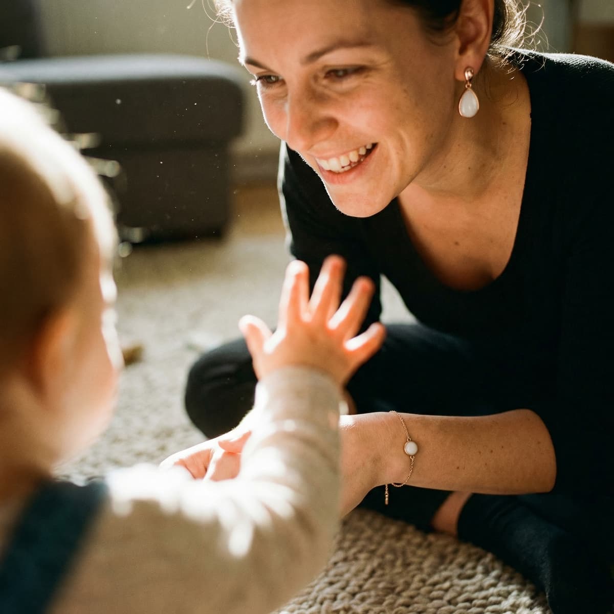 Smiling mother plays with her baby on a sunlit rug, wearing a white teardrop breastmilk keepsake earring and matching bracelet, a meaningful 1 year breastfeeding gift.