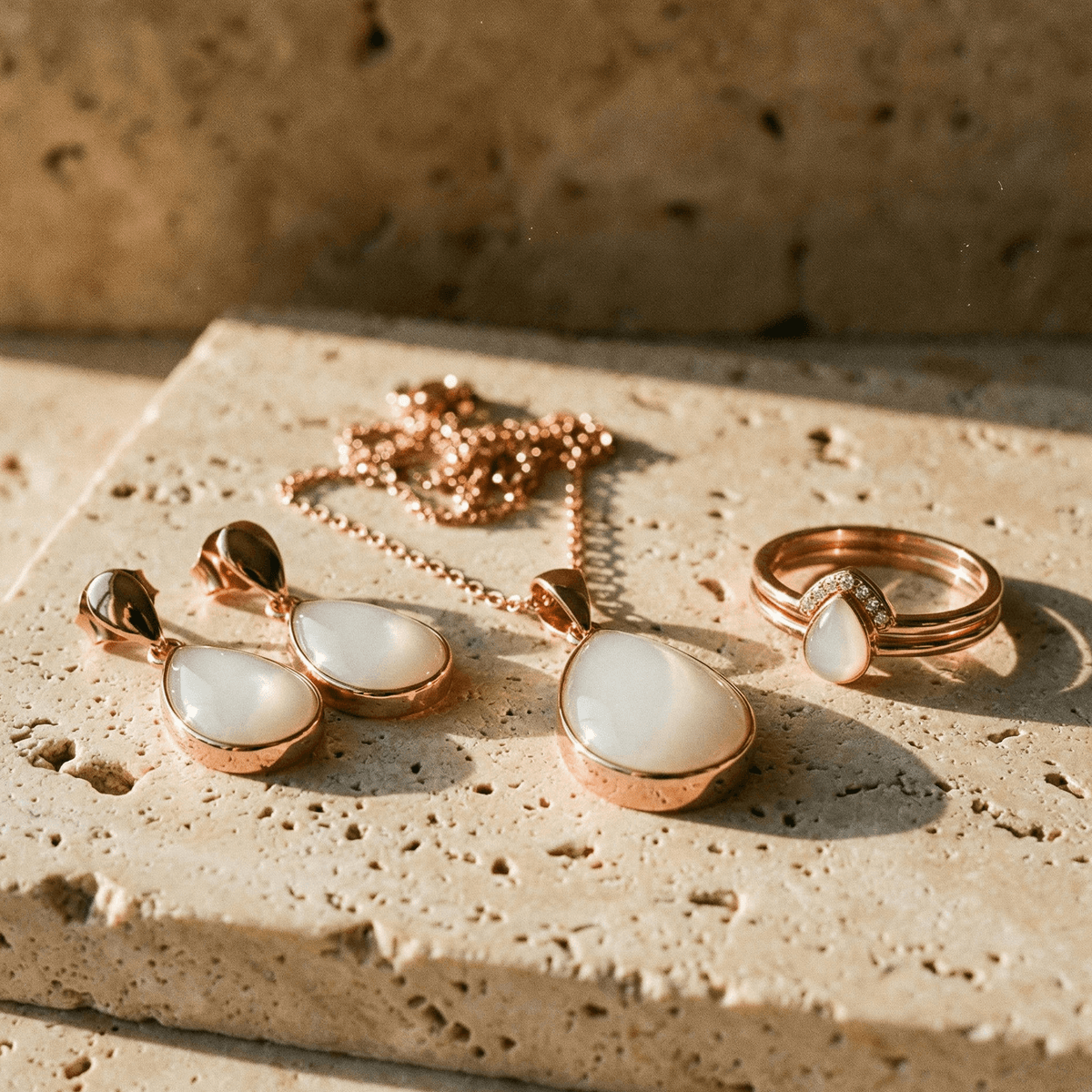 Rose gold breastmilk keepsake jewelry set with teardrop pendant necklace, matching earrings, and ring featuring milky white stones, a thoughtful 6 months breastfeeding award gift on a beige stone surface in warm light.