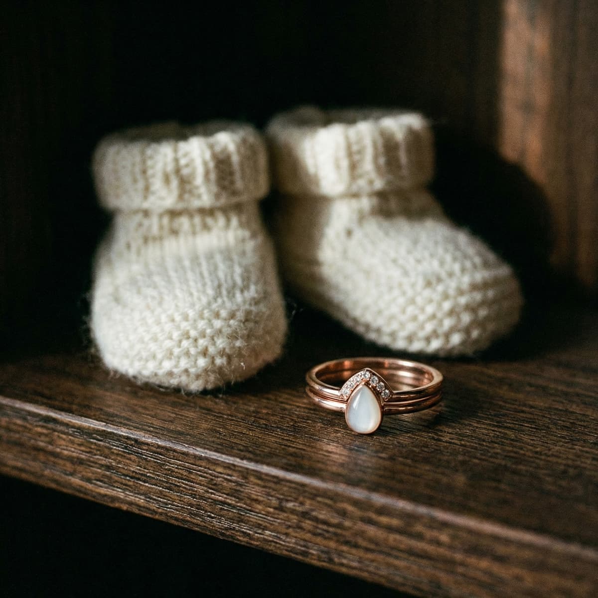 Cozy baby booties on a rustic wooden shelf beside a rose-gold ring with a milky teardrop stone, a sentimental baby keepsake for mom.