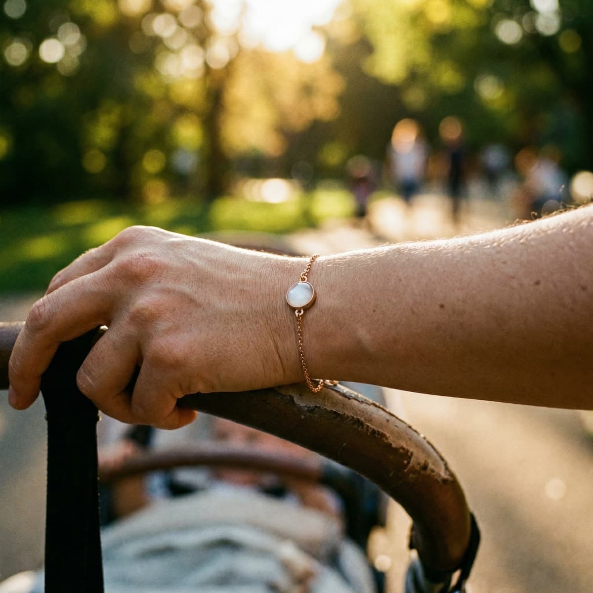 Close-up of a mom’s hand pushing a stroller, wearing a delicate gold bracelet with a milky white resin stone as a meaningful baby keepsake for mom in warm outdoor light.
