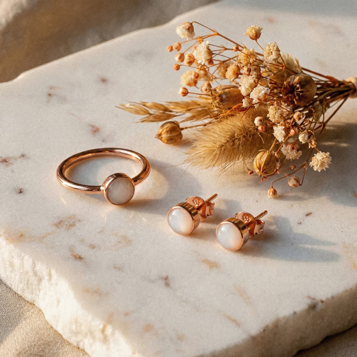 Rose-gold ring and matching stud earrings with milky white stones on a marble slab beside dried flowers, a timeless idea for the best push presents for new moms.