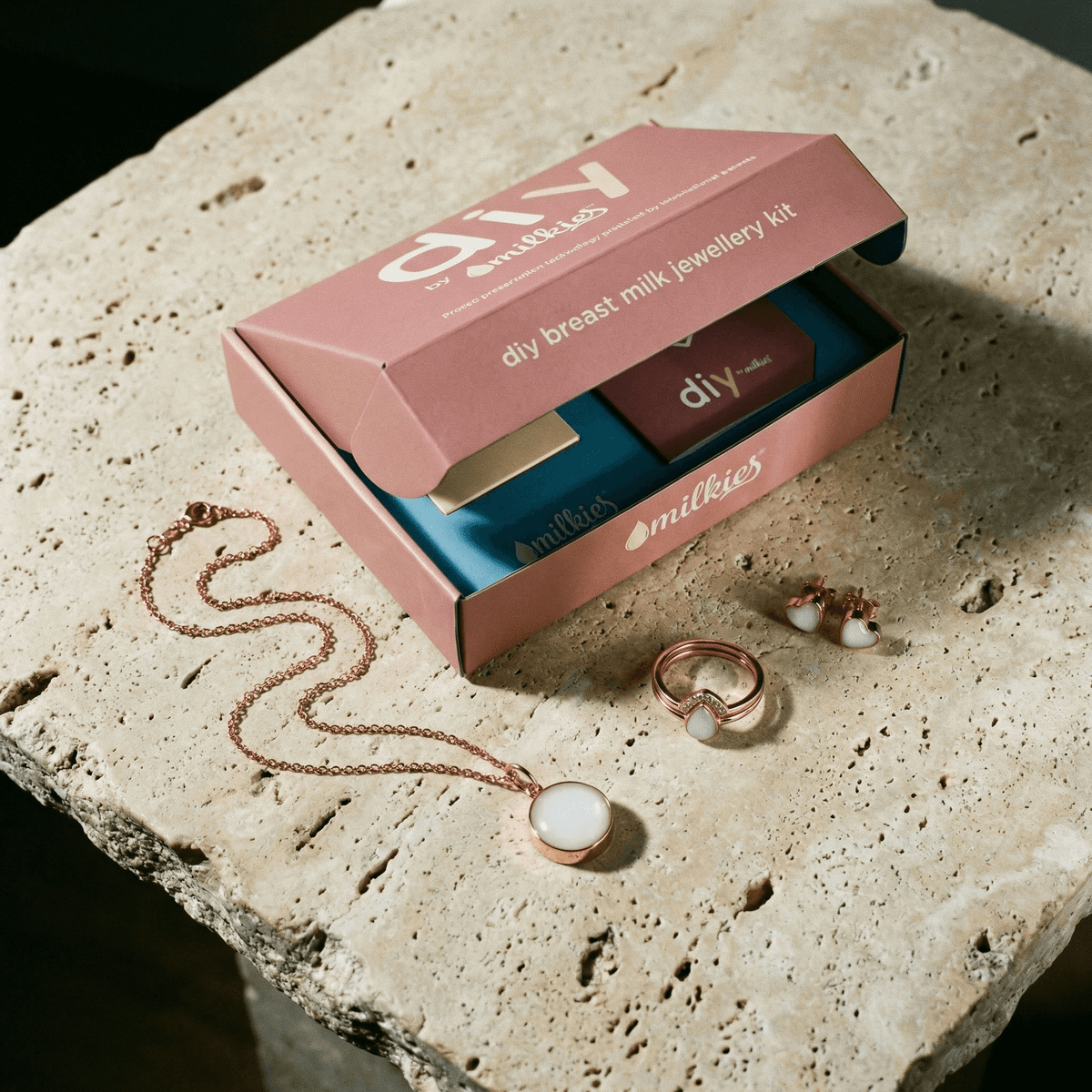 Open DIY by MILKIES breast milk jewellery diy kit in a pink box on a stone surface, showing rose-gold pendant necklace, ring, and stud earrings with milky white resin breastmilk keepsakes.