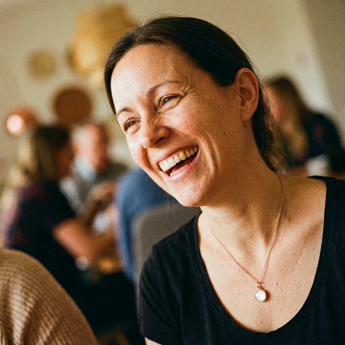 Smiling mother in warm candid light wearing a delicate necklace with a round white breastmilk keepsake pendant, a meaningful breastfeeding celebration moment.