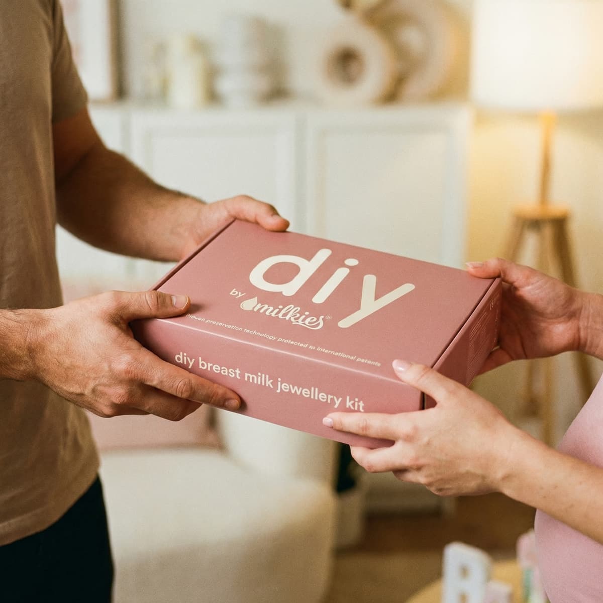 Hands exchange a pink DIY by MILKIES breast milk jewellery kit box, a thoughtful breastfeeding gift for making a keepsake at home.