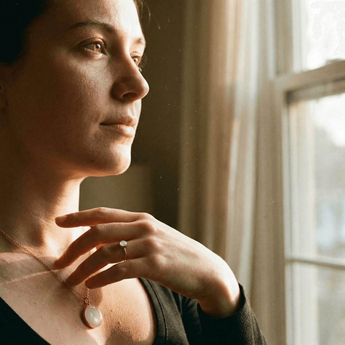 Woman gazing out a sunlit window wearing a delicate breastmilk resin pendant necklace, a meaningful breastfeeding journey gift for new moms.