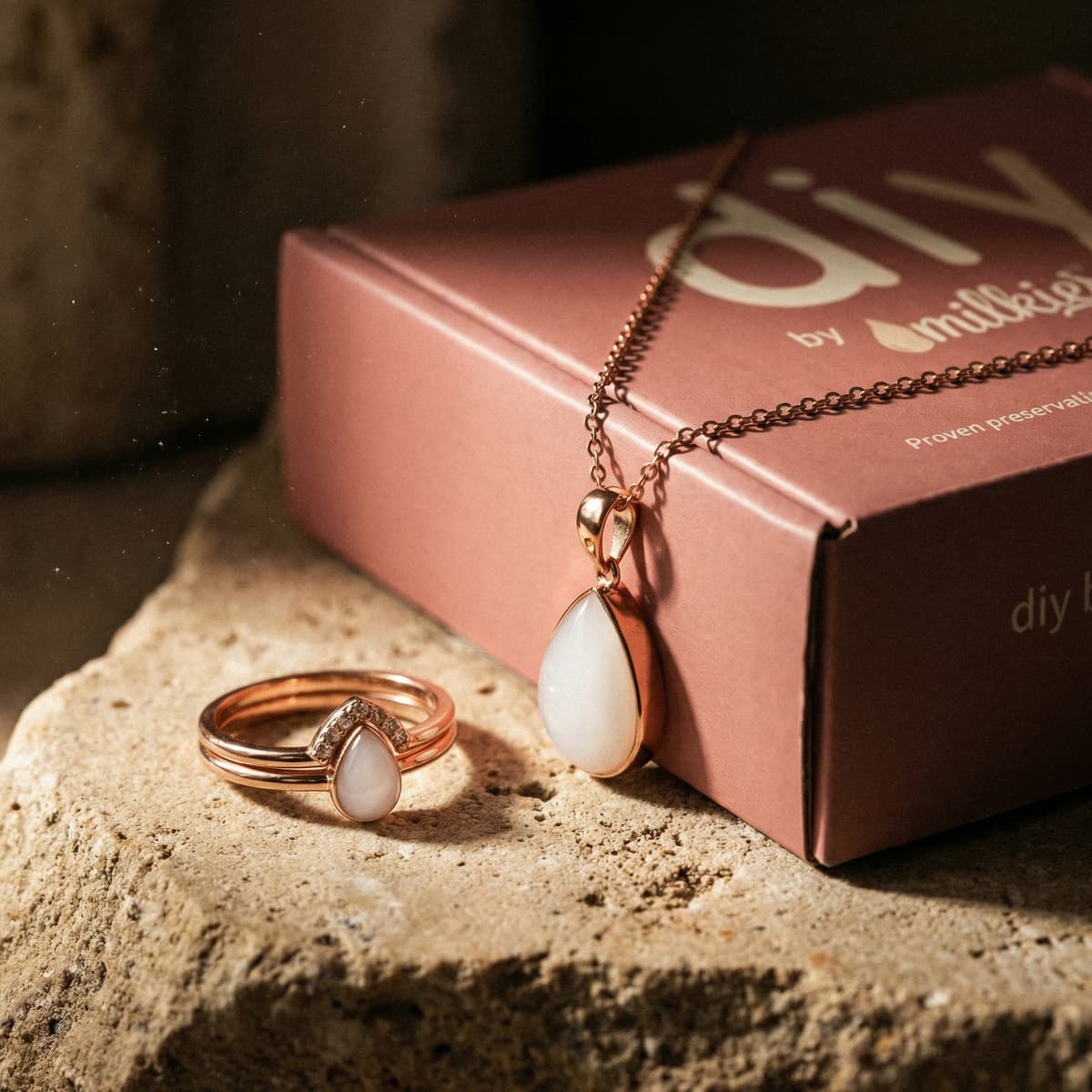 Rose-gold breastmilk keepsake necklace and ring with milky-white teardrop stones beside a pink DIY by MILKIES kit box, a meaningful breastfeeding journey gift for moms