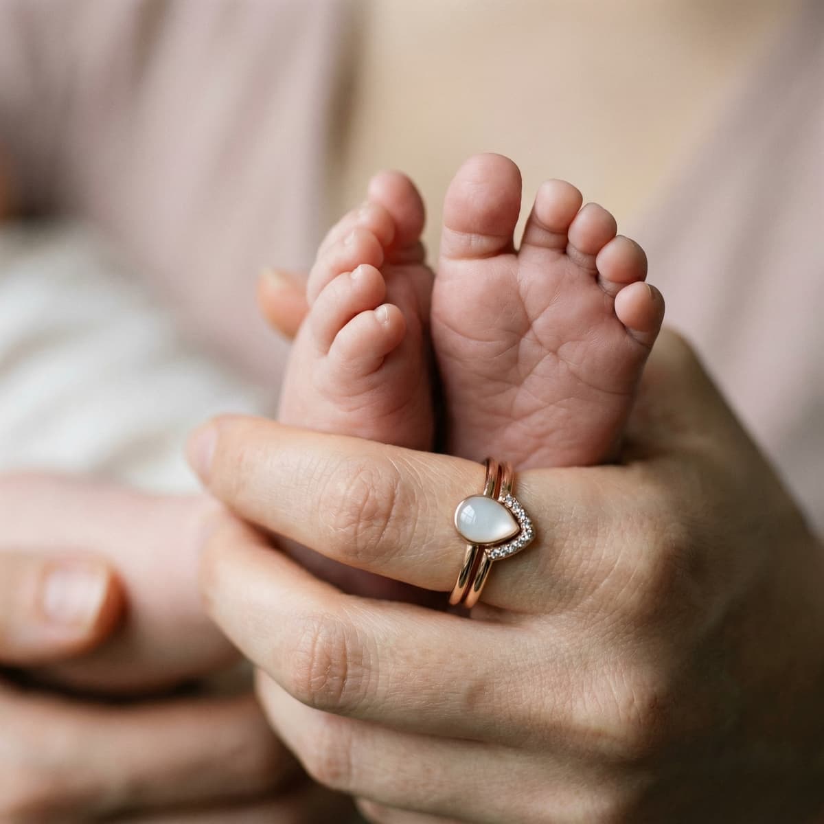 Mother holds baby’s feet while wearing a heart-shaped ring with a milky white stone, a rose-gold toned breastfeeding keepsake jewelry piece.