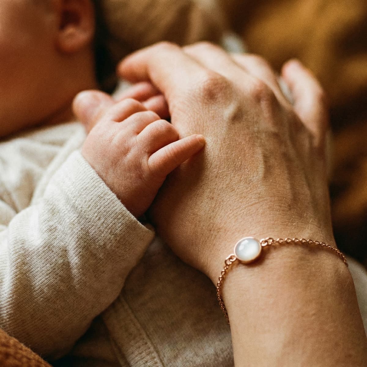 Mother holding a baby’s hand while wearing a delicate keepsake breastmilk bracelet featuring a milky-white resin charm on a rose-gold chain.