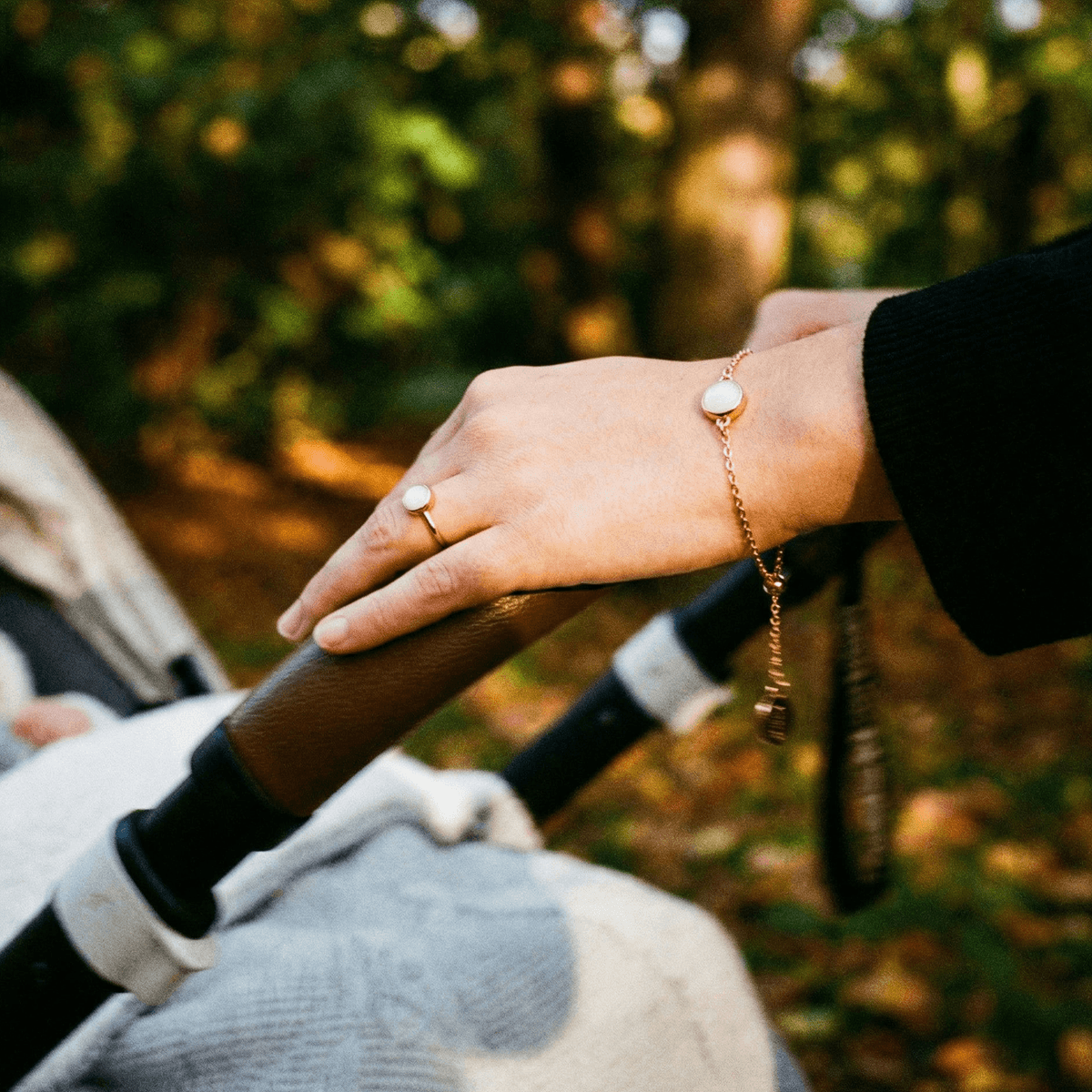 Hand pushing a stroller outdoors while wearing a rose-gold keepsake bracelet and ring with a white stone, showcasing a breastmilk bracelet diy kit jewelry design.