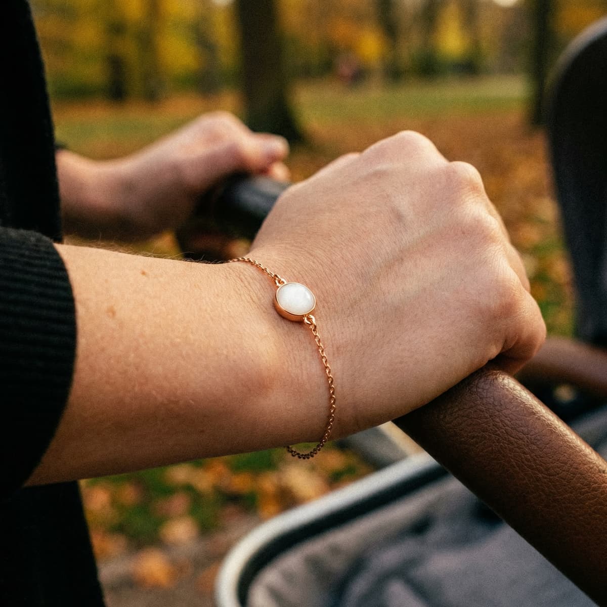 Rose gold bracelet with a round white resin stone charm from a breastmilk bracelet kit, worn on a mother’s wrist as she pushes a stroller outdoors.