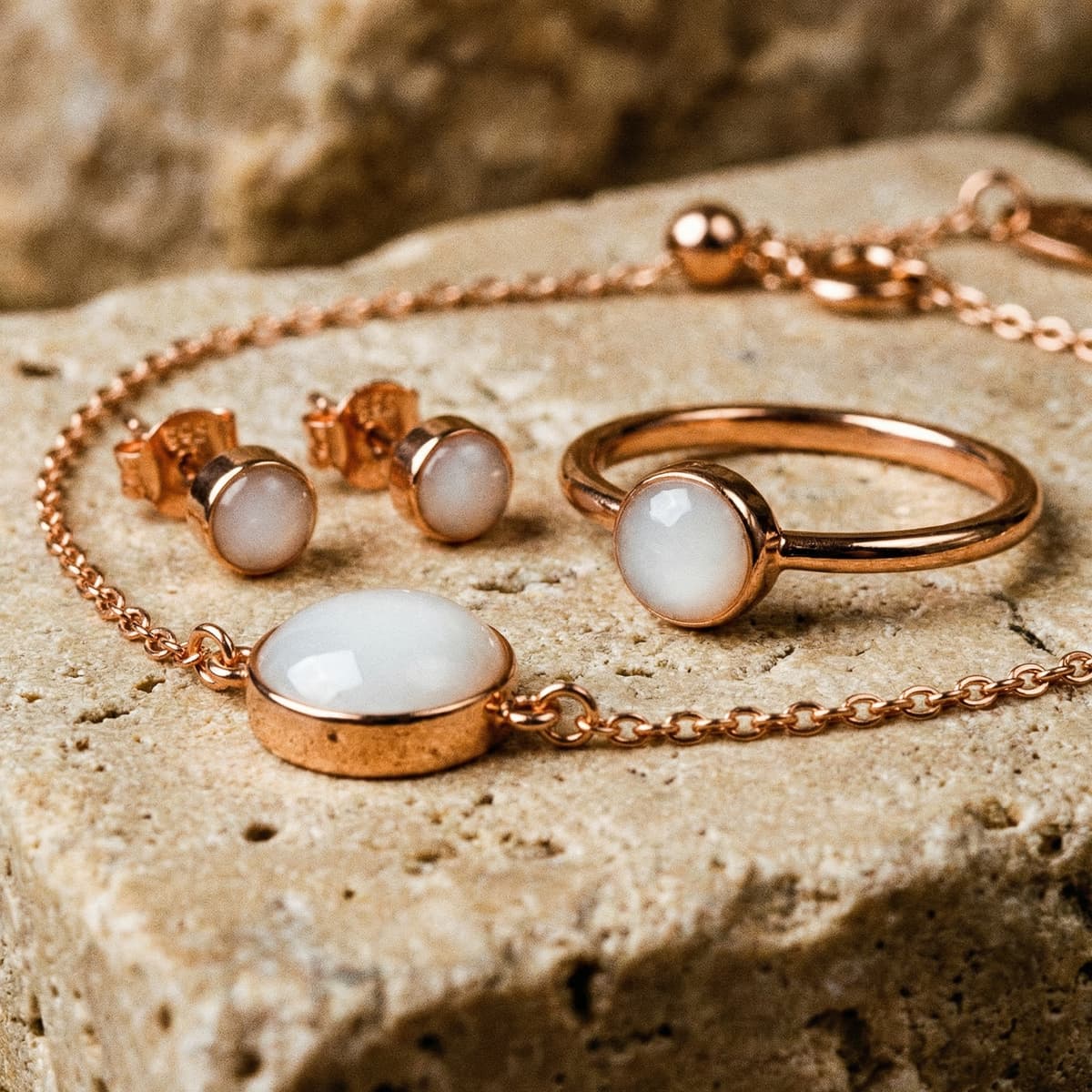 Rose gold breastmilk bracelet kit keepsake set with a delicate chain bracelet featuring a milky white stone, matching stud earrings and ring displayed on a textured stone background