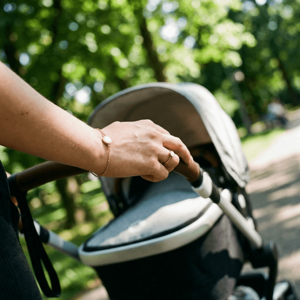 Mother outdoors pushing a stroller while wearing a delicate keepsake bracelet made with a breastmilk bracelets kit, featuring an oval white stone on a gold chain.