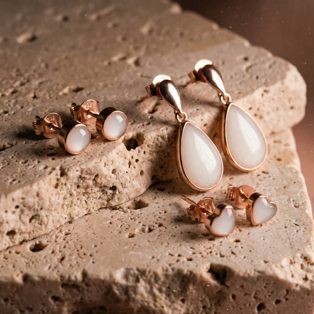 Rose gold keepsake jewelry display featuring a breastmilk earring set with milky white resin stones, including teardrop dangle earrings, round stud earrings, and small heart studs on a textured stone surface.