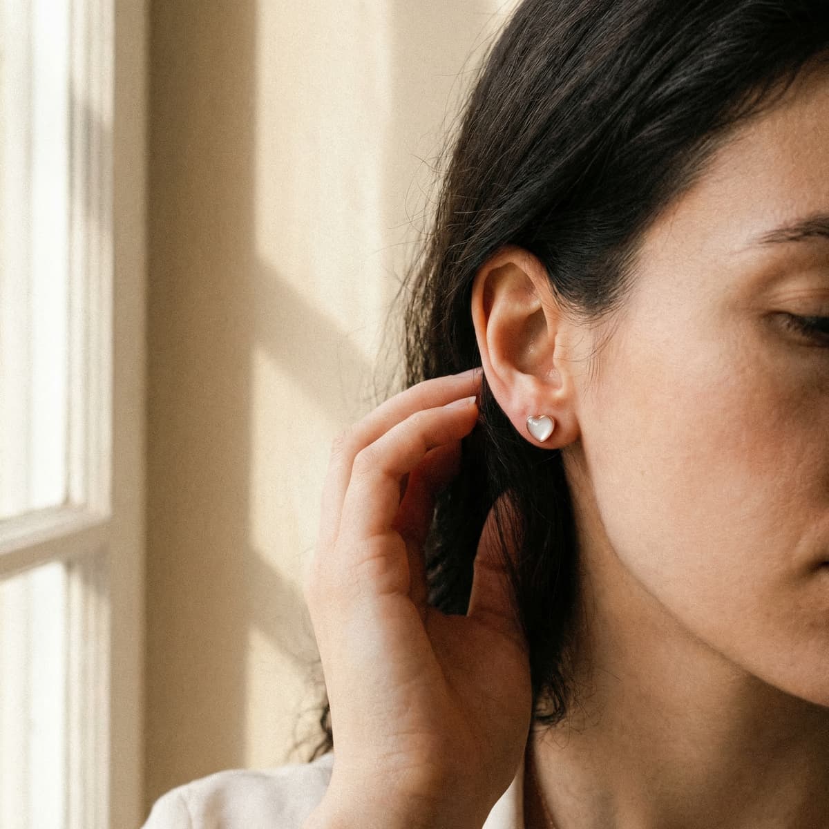 Close-up of a woman wearing a small heart-shaped stud made with a breastmilk earring kit, photographed by a sunlit window.
