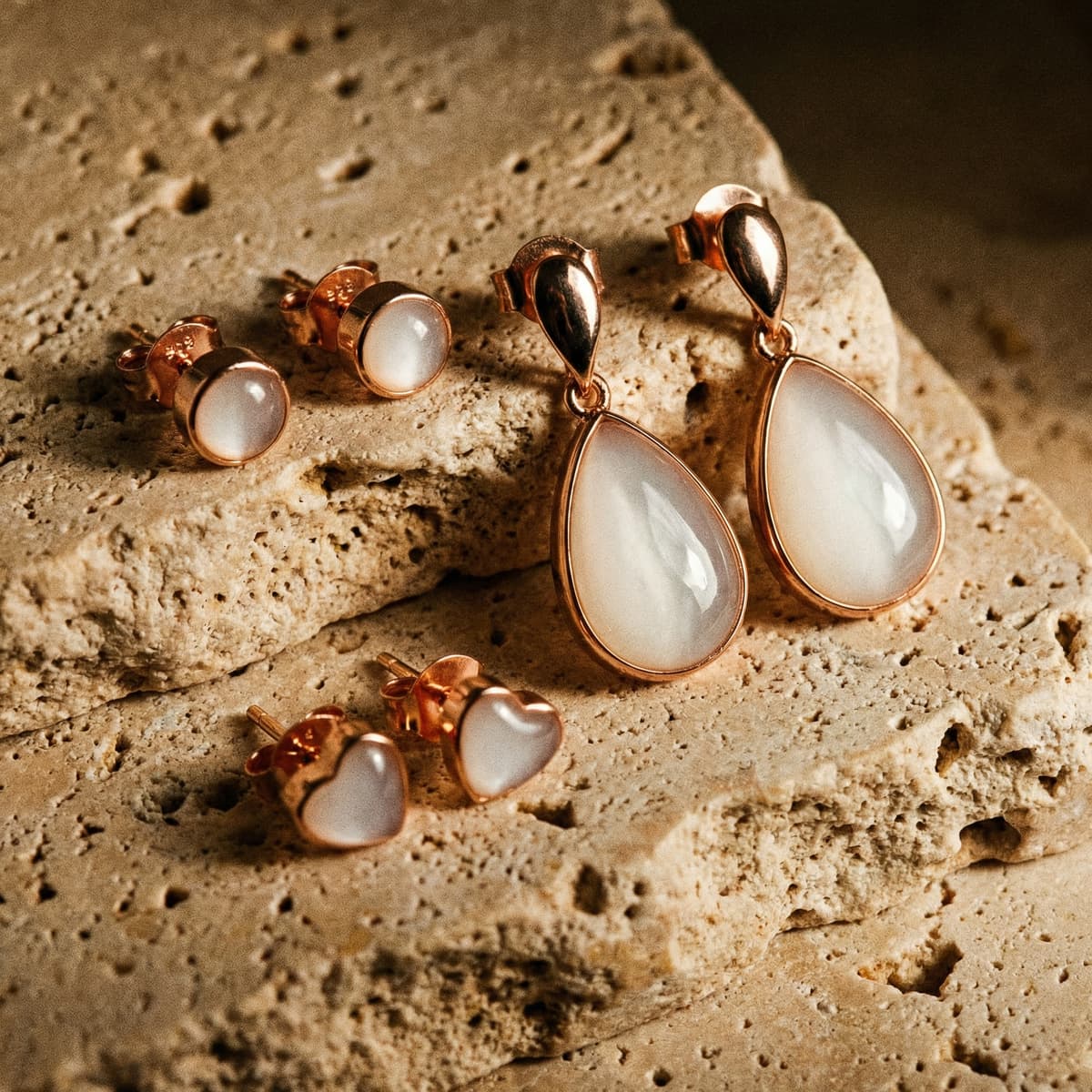 Rose gold keepsake earrings from an at-home breastmilk earring kit displayed on textured stone, featuring milky white resin teardrop drops with matching round and heart stud settings.