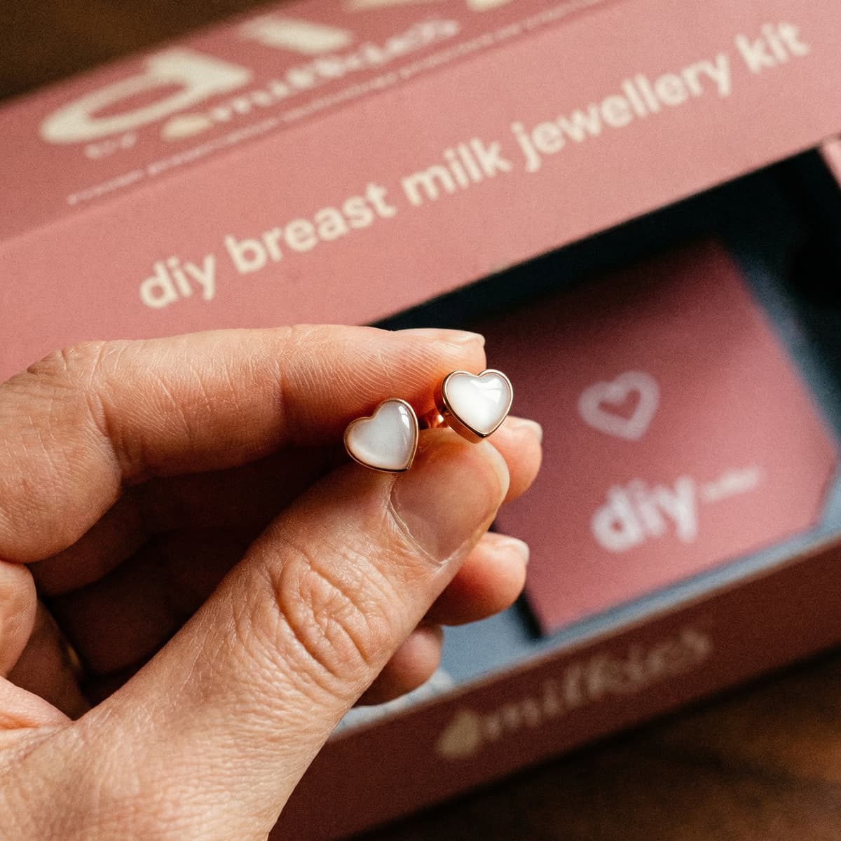 Hand holding heart-shaped resin breastmilk earrings in gold-toned settings, with the DIY by MILKIES breast milk jewellery kit box blurred in the background.