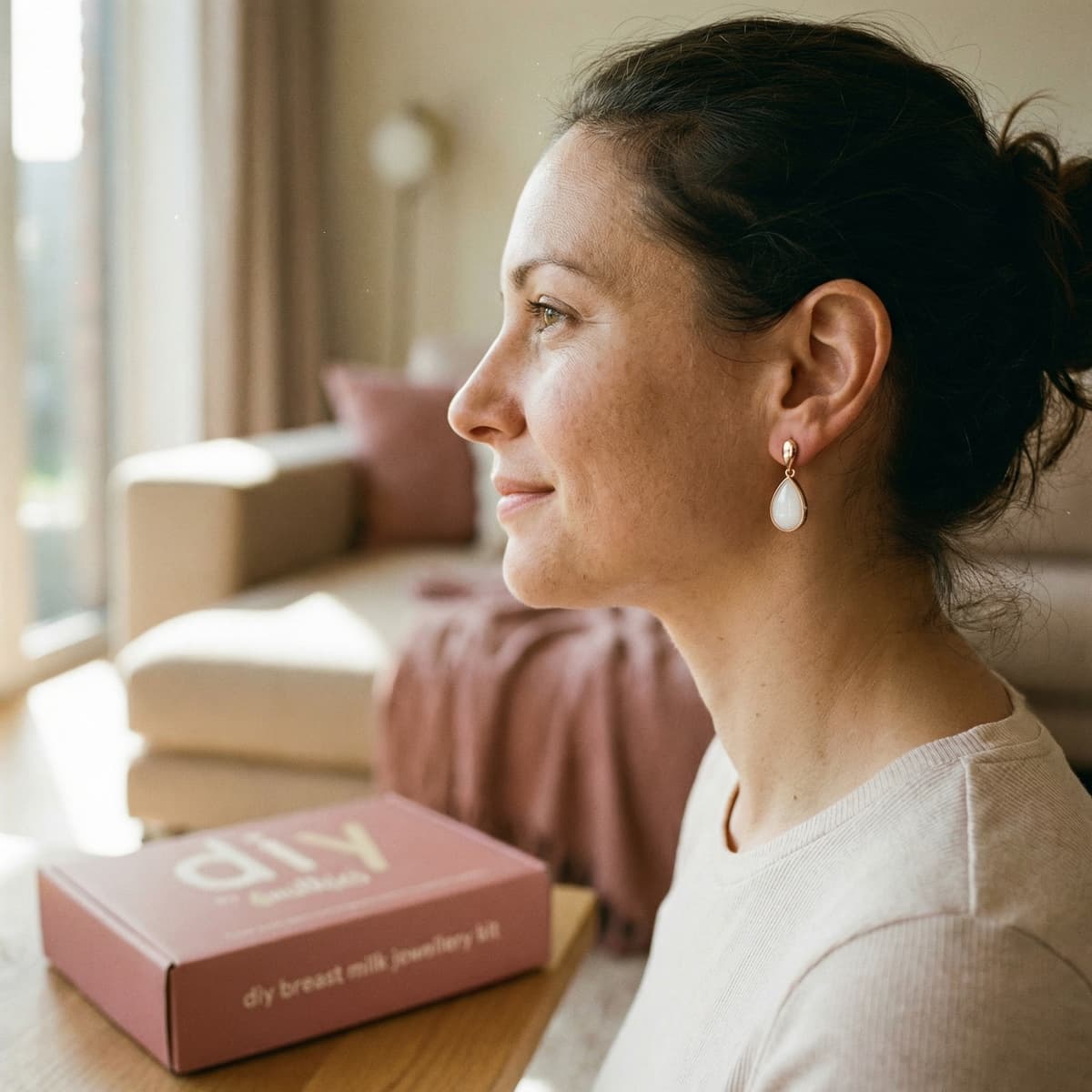 Woman wearing elegant breastmilk earrings by a window at home beside the DIY by MILKIES breastmilk jewellery kit box, highlighting why creating a personal keepsake yourself is a meaningful choice.