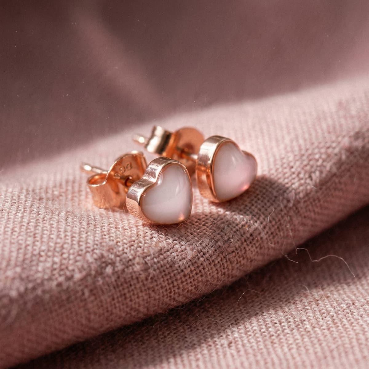 Heart-shaped rose gold stud earrings with milky white resin, showing a DIY keepsake made with a breastmilk earrings kit on soft textured fabric.