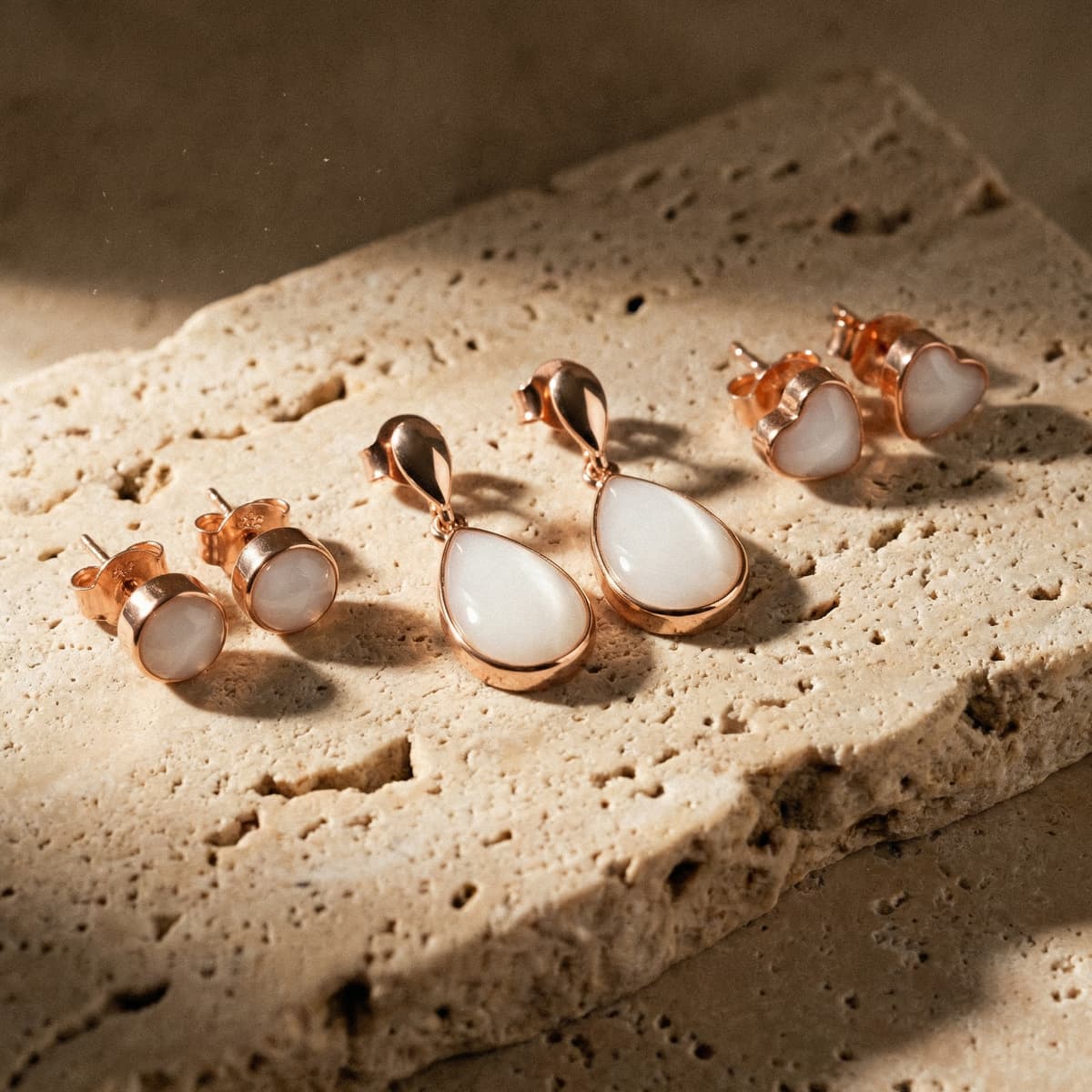 Rose-gold breastmilk earrings in heart, round stud, and teardrop dangle settings made with a breastmilk earrings making kit, showing milky-white resin keepsakes on a textured stone surface.