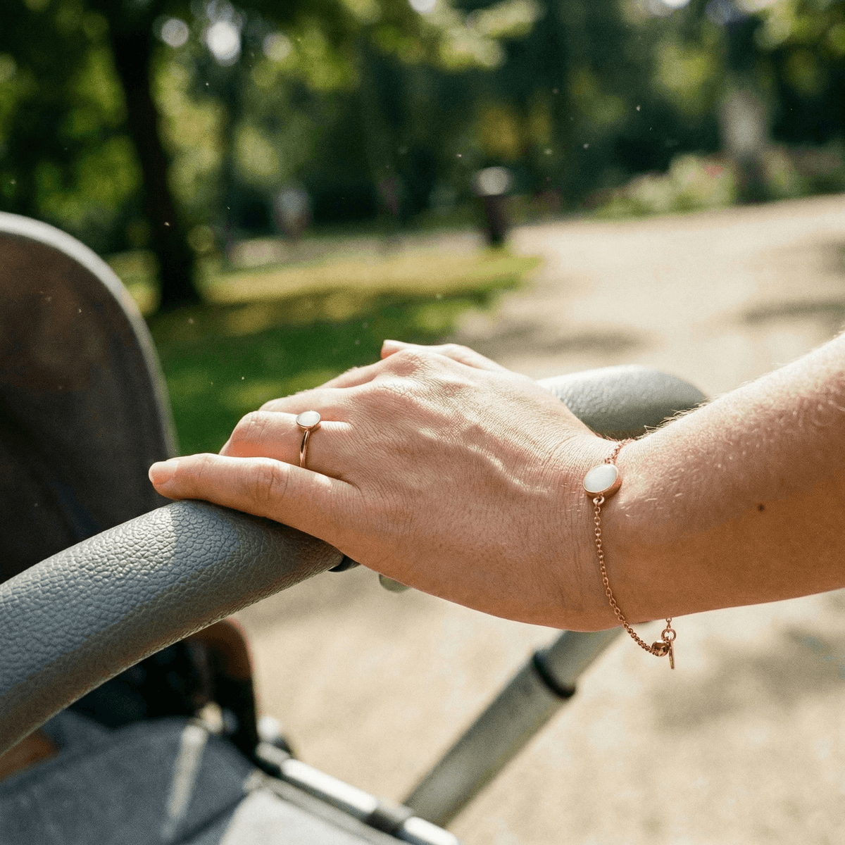 Hand resting on an outdoor stroller handle wearing a gold ring and matching bracelet with milky-white stones, showcasing elegant breastmilk jewellery keepsakes.