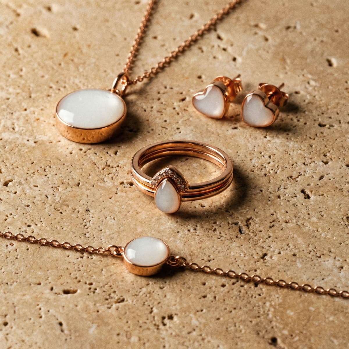 Rose-gold keepsake set made with a breastmilk jewelry making kit, showing a milky white pendant necklace, bracelet, stacking ring with teardrop stone, and heart stud earrings on a textured stone background.