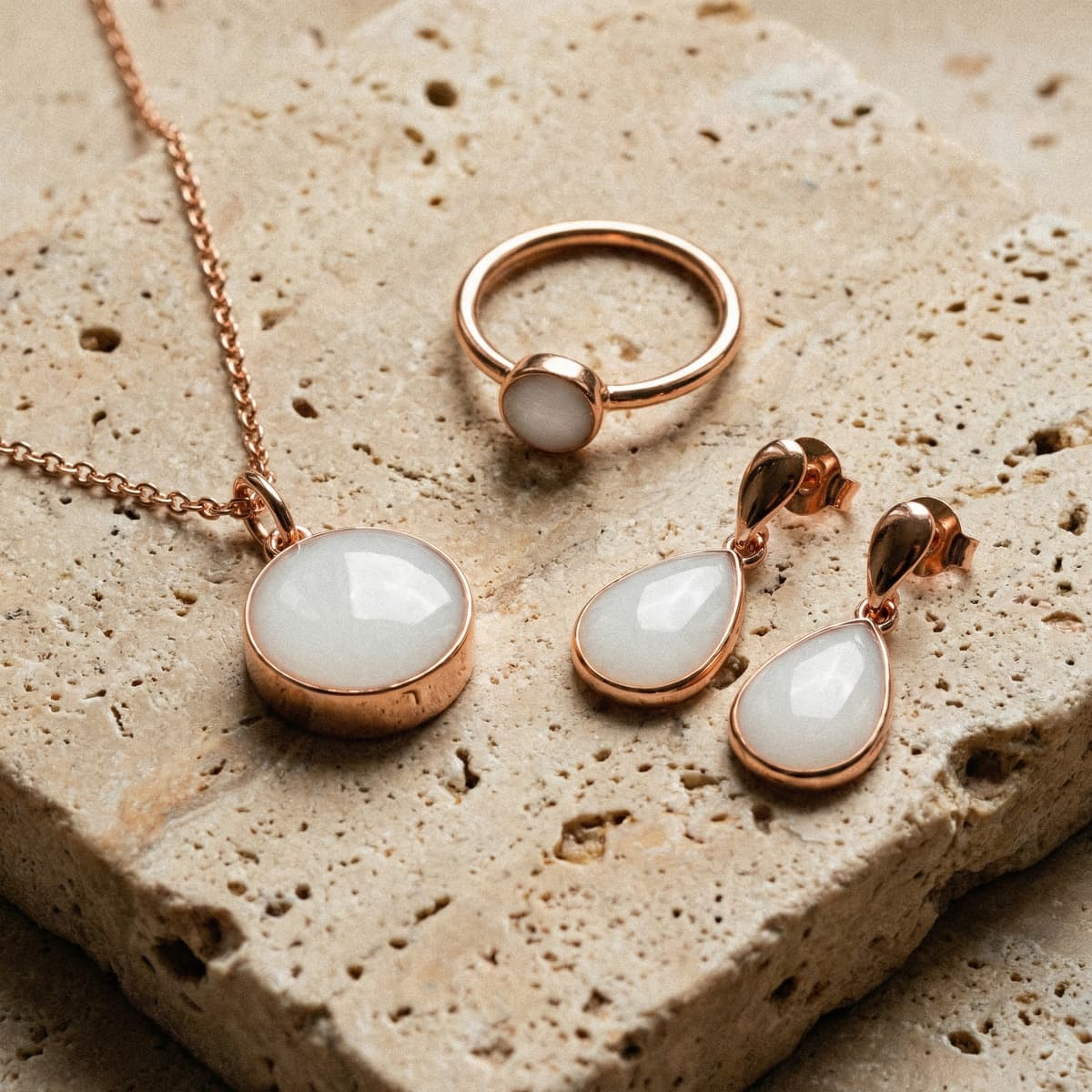 Rose gold necklace, ring, and teardrop earrings with milky white stones, showcasing keepsakes made using a breastmilk jewelry resin kit on a textured beige stone surface.