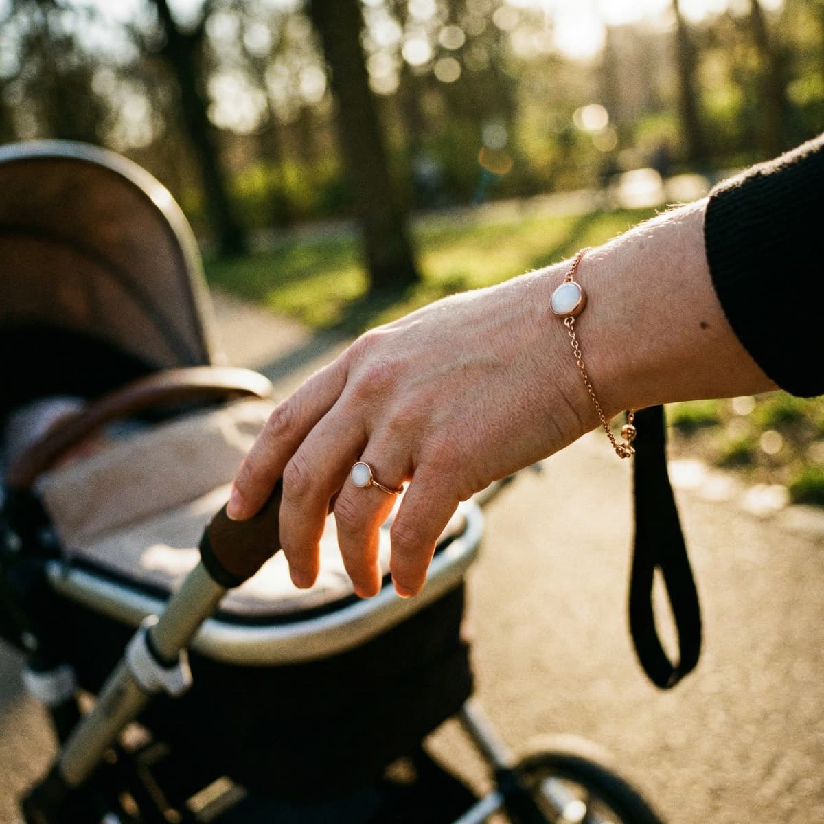 Mother’s hand pushing a stroller outdoors in warm sunlight, wearing a delicate gold bracelet and ring with a milky-white breastmilk keepsake stone.