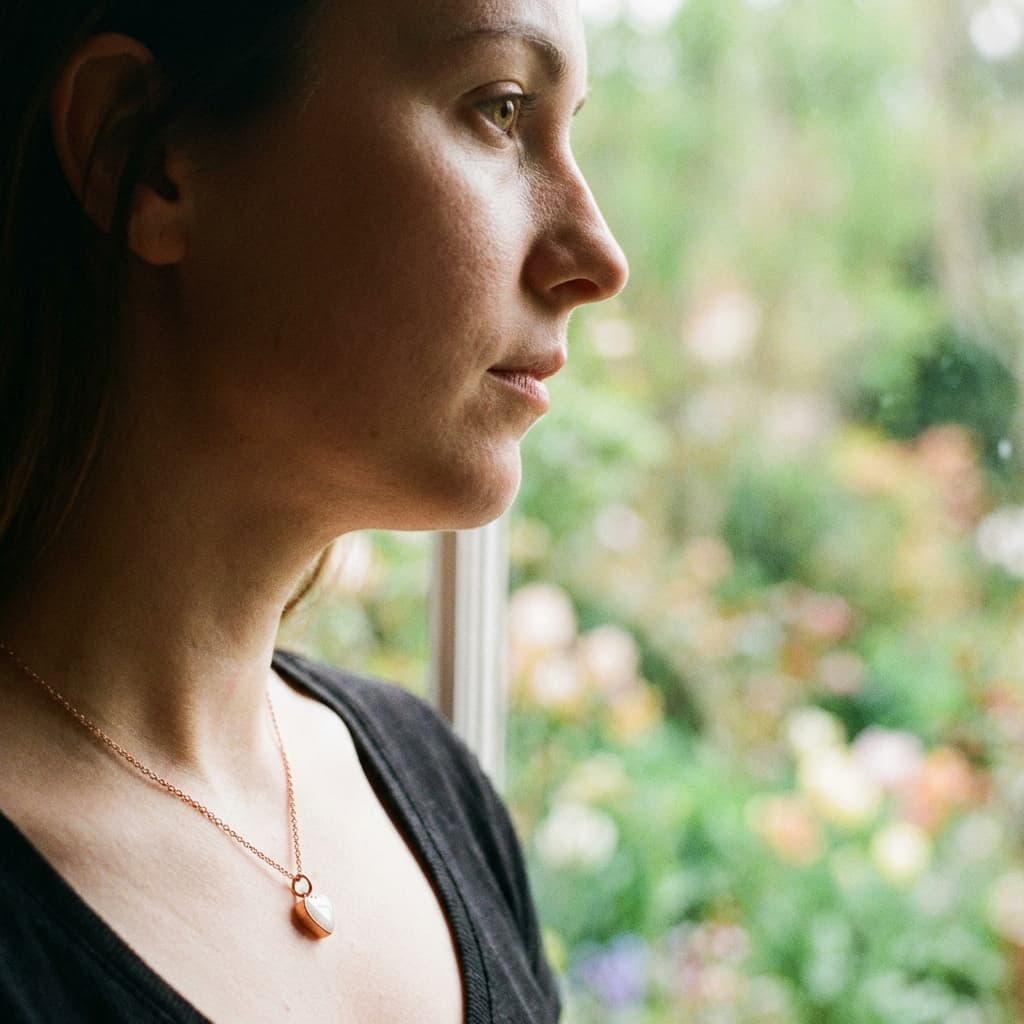 Mother wearing a delicate breastmilk keepsake pendant made with a breastmilk necklace diy kit, standing by a window and looking outside.