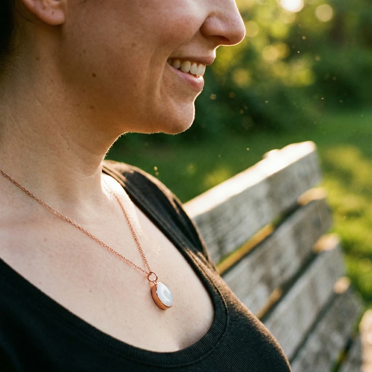 Smiling woman outdoors wearing a rose-gold pendant necklace made with a breastmilk necklace kit, featuring a milky white oval keepsake charm on a delicate chain.