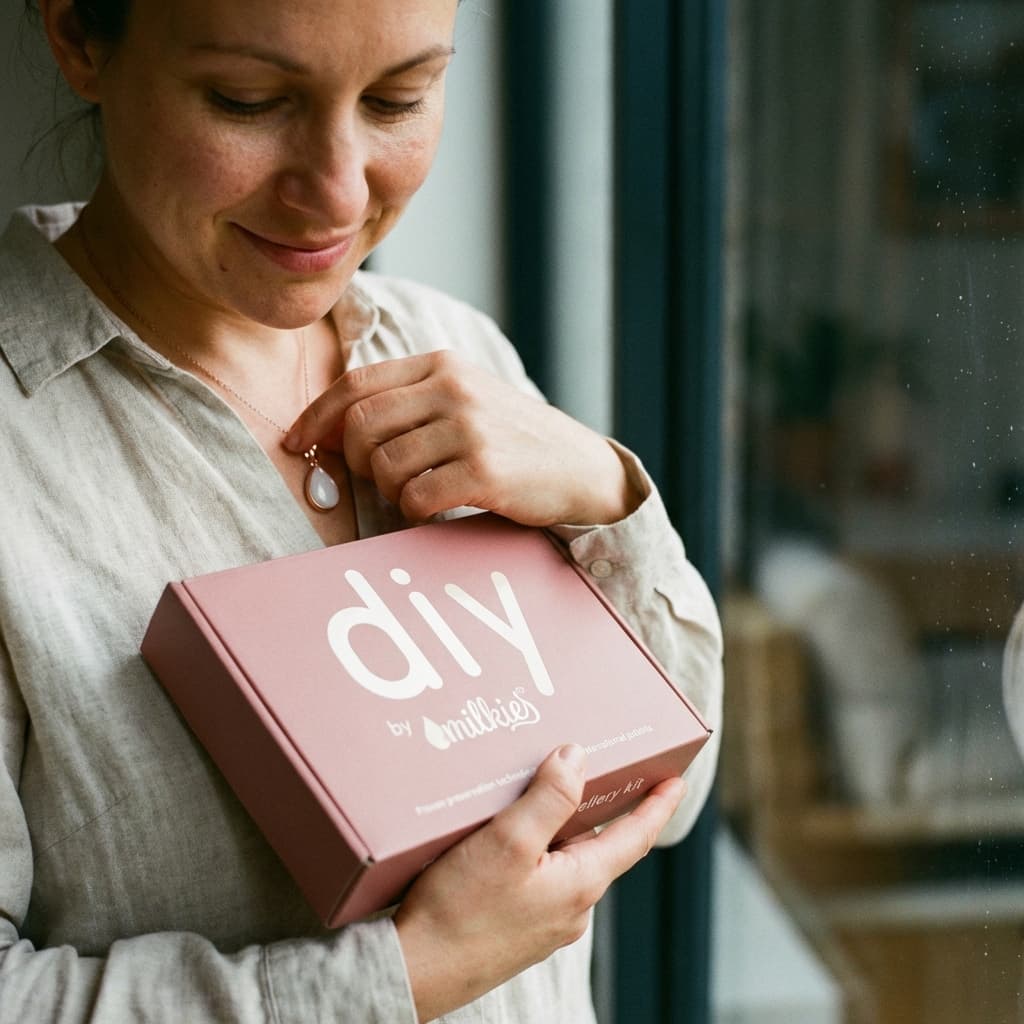 Mother holds a pink DIY by MILKIES box and touches a keepsake pendant, showing why a breastmilk necklace kit is a meaningful, private at-home way to create breastmilk jewelry.