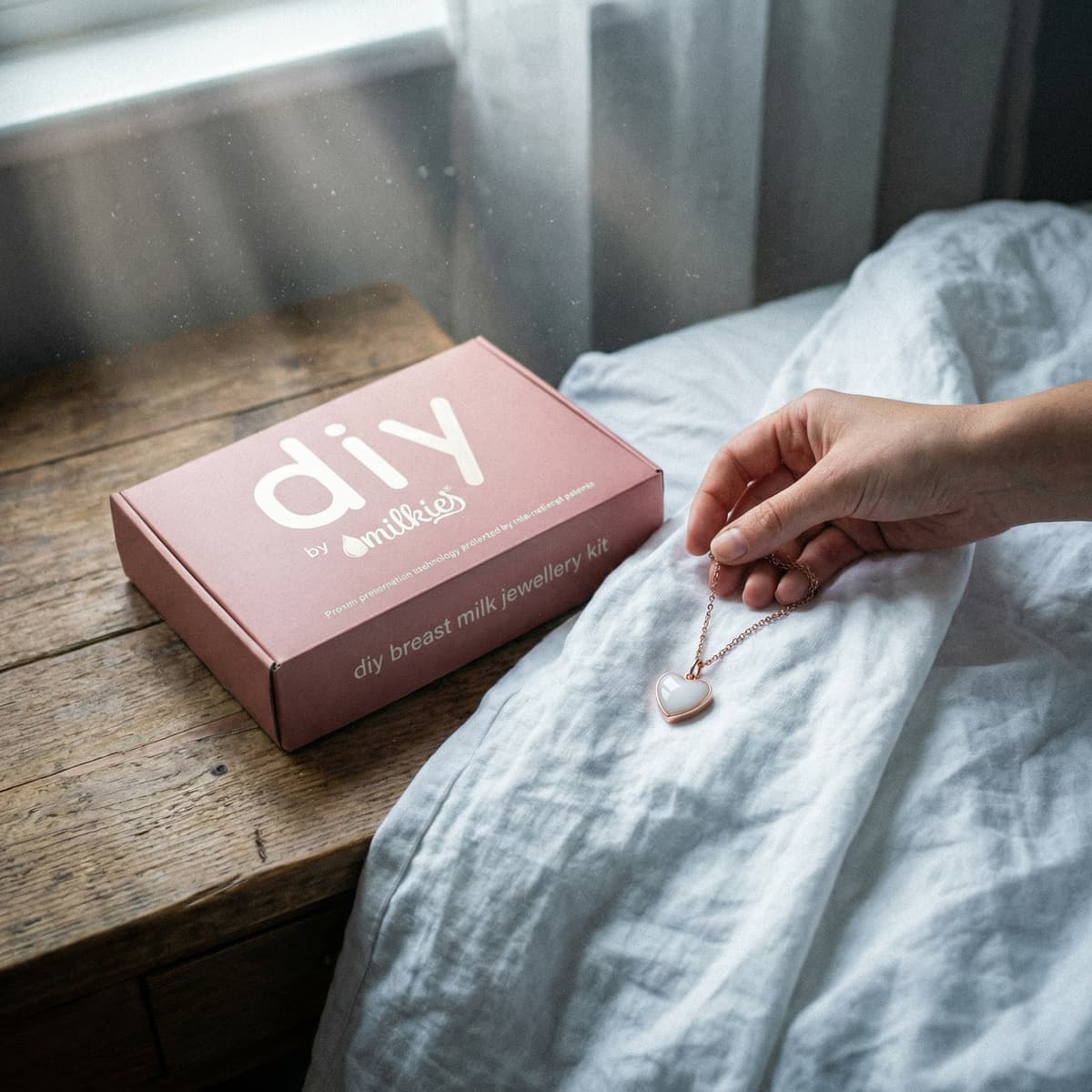 Hand holding a rose-gold heart pendant from an at-home DIY kit, showing breastmilk necklaces beside the DIY by MILKIES breast milk jewellery kit box on a wooden bedside table.