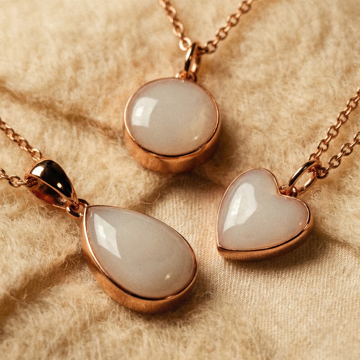 Close-up of three rose-gold breastmilk necklaces featuring milky white resin pendants in round, teardrop, and heart shapes displayed on soft beige fabric.