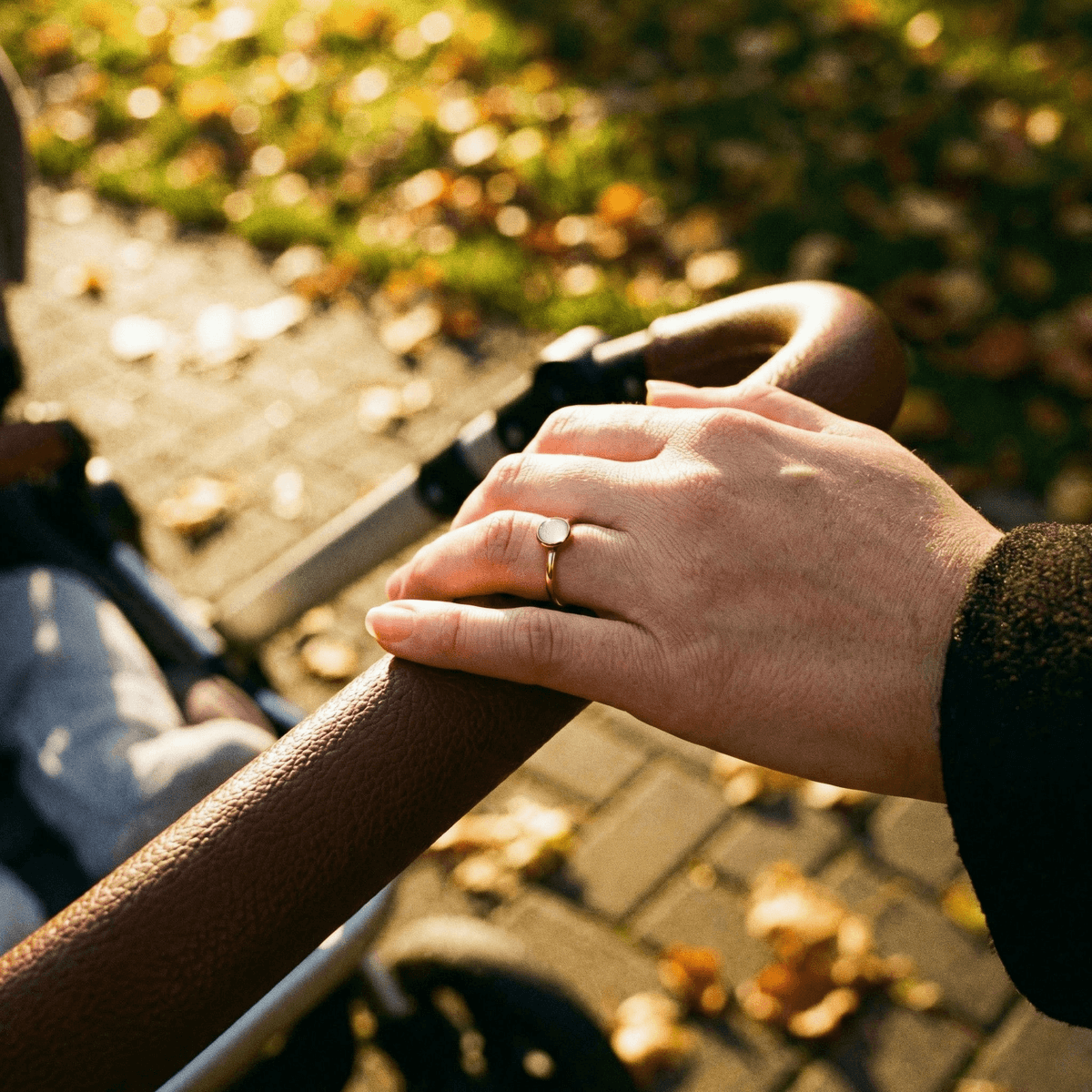 A minimalist gold ring with a small milky-white stone, a breastmilk ring worn on a woman’s hand resting on a stroller handle in warm outdoor light.