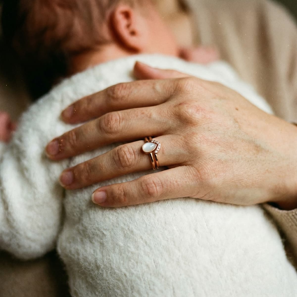 Mother cuddling a newborn while wearing a rose-gold breastmilk ring with a milky white stone, illustrating why a DIY by MILKIES at-home keepsake kit is a meaningful, private way to preserve breastfeeding memories.