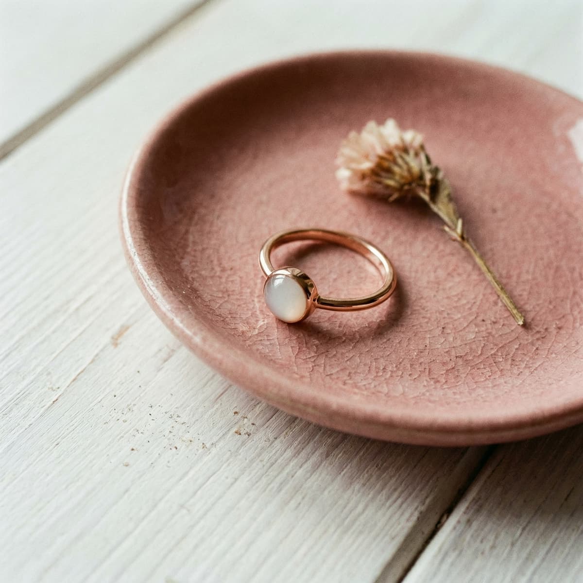 Delicate gold ring with a milky white stone on a pink ceramic dish beside a dried flower, showcasing a DIY breastmilk ring kit keepsake style.