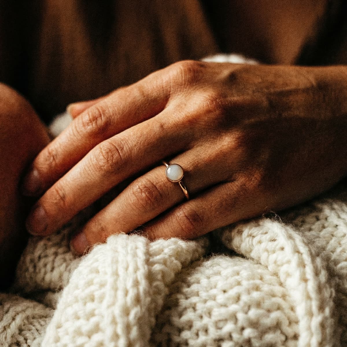 Mother’s hand resting on a cozy knit blanket, wearing a delicate gold keepsake ring with a milky white stone made using a breastmilk ring making kit.