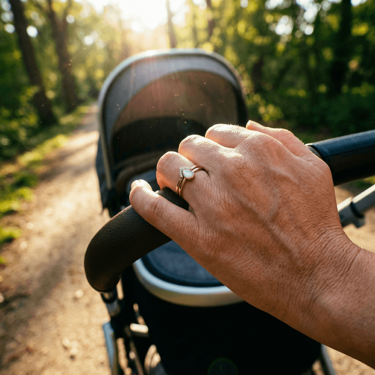 Hand wearing elegant breastmilk rings with a milky teardrop stone and pavé halo, resting on a baby stroller handle during a sunlit walk in the park