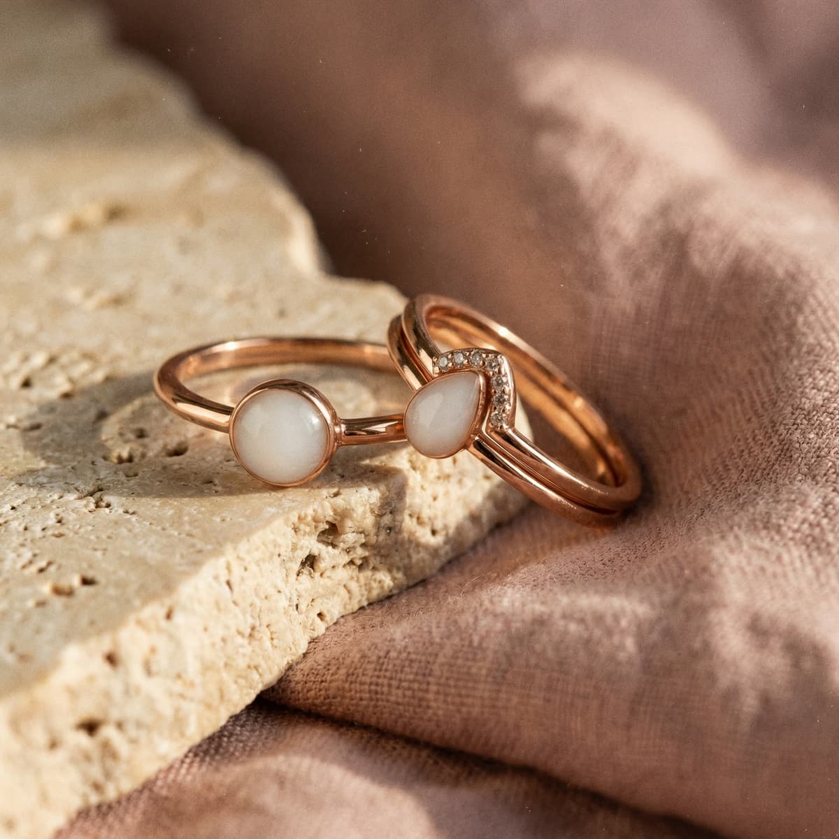Rose gold keepsake breastmilk rings featuring milky white round and teardrop stones with crystal accents, displayed on a textured stone over soft fabric.