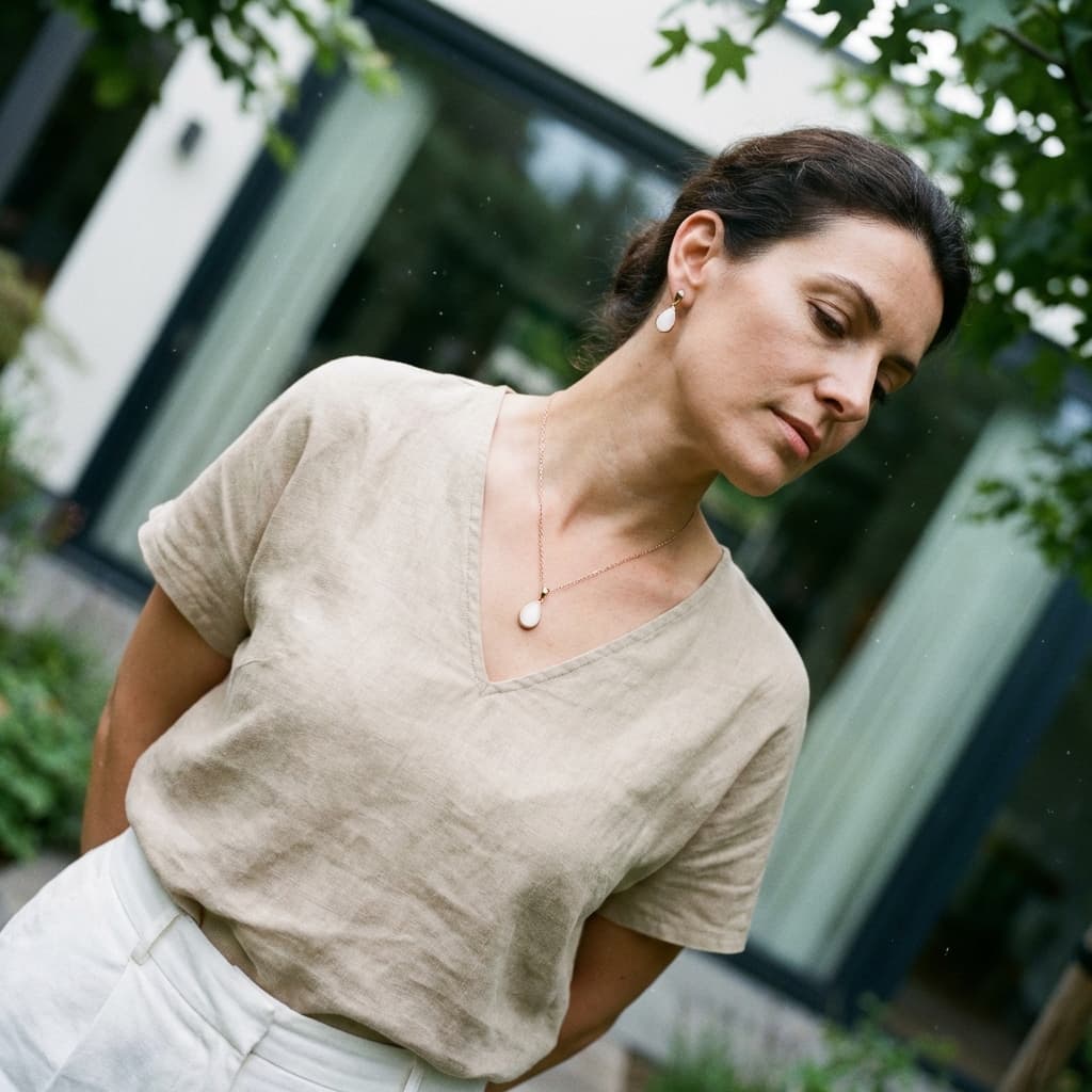 Woman outdoors wearing DIY by MILKIES diy breast milk jewellery—a delicate gold necklace and matching earrings with milky white resin stones—paired with a beige top.