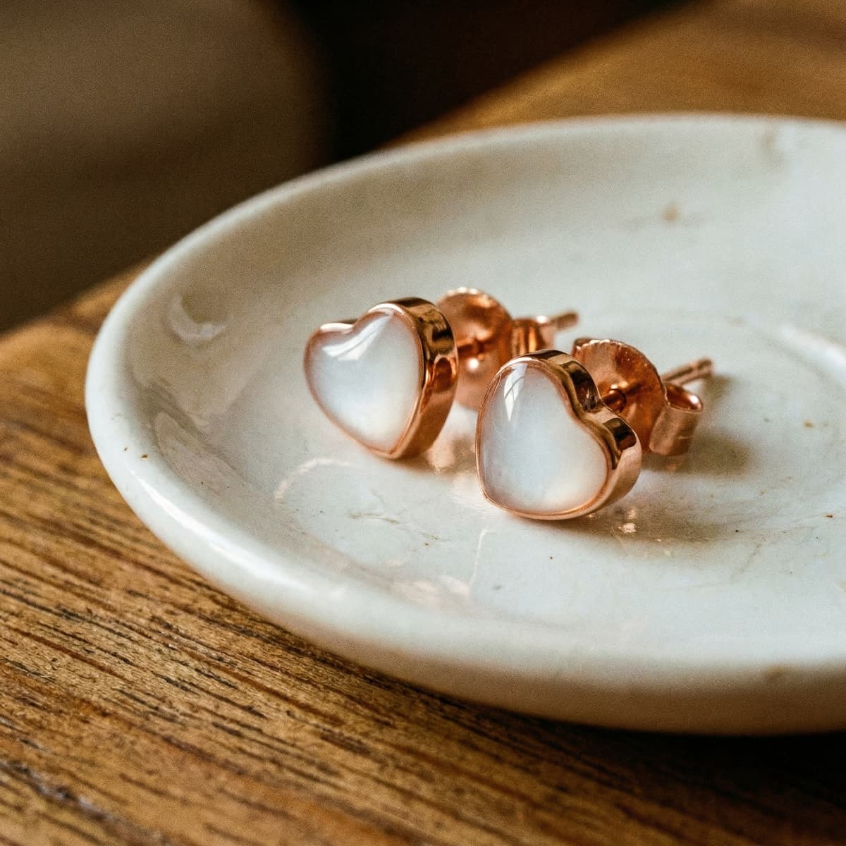 Rose-gold heart-shaped stud earrings with milky white resin inlays, a diy breastmilk earring keepsake pair displayed on a small ceramic dish on a wooden surface.