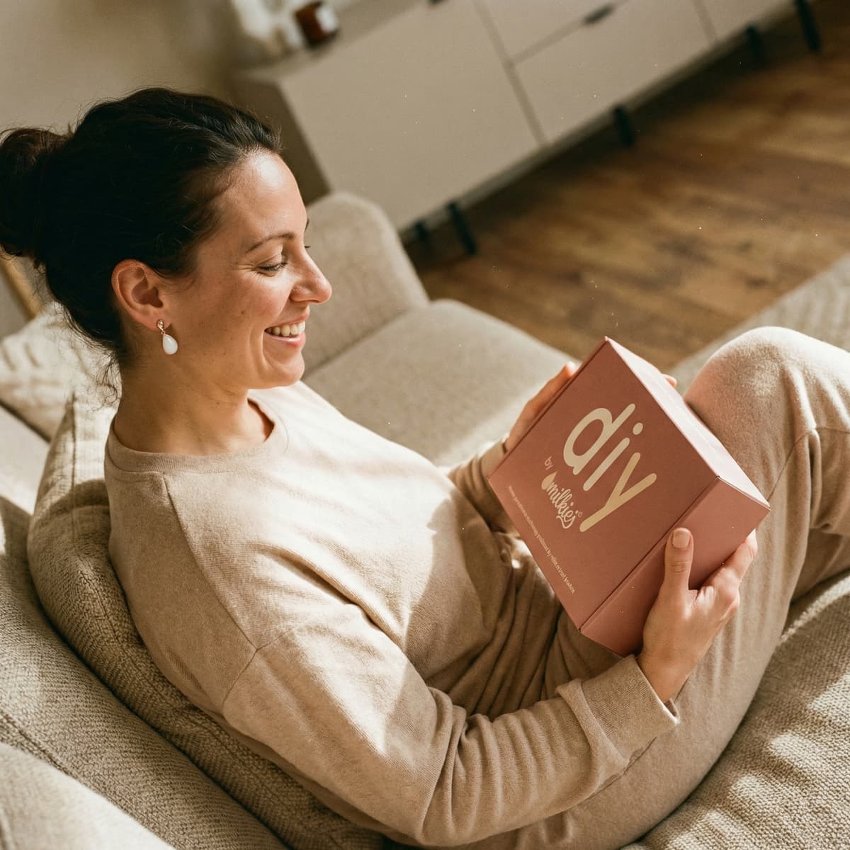 Smiling mom relaxing on a sofa holding a DIY by MILKIES kit box, showing why making a diy breastmilk earring at home is a meaningful and private keepsake option.