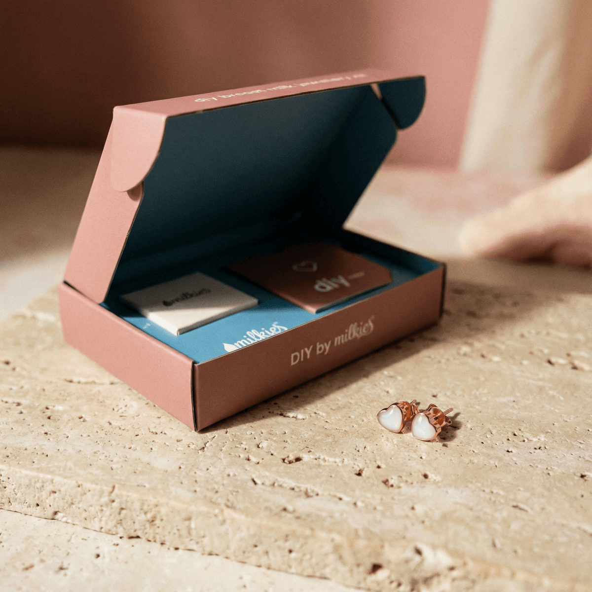 Open DIY by MILKIES box with tools and instructions beside rose-gold heart stud settings, showcasing a diy breastmilk earring kit for making keepsake earrings at home.
