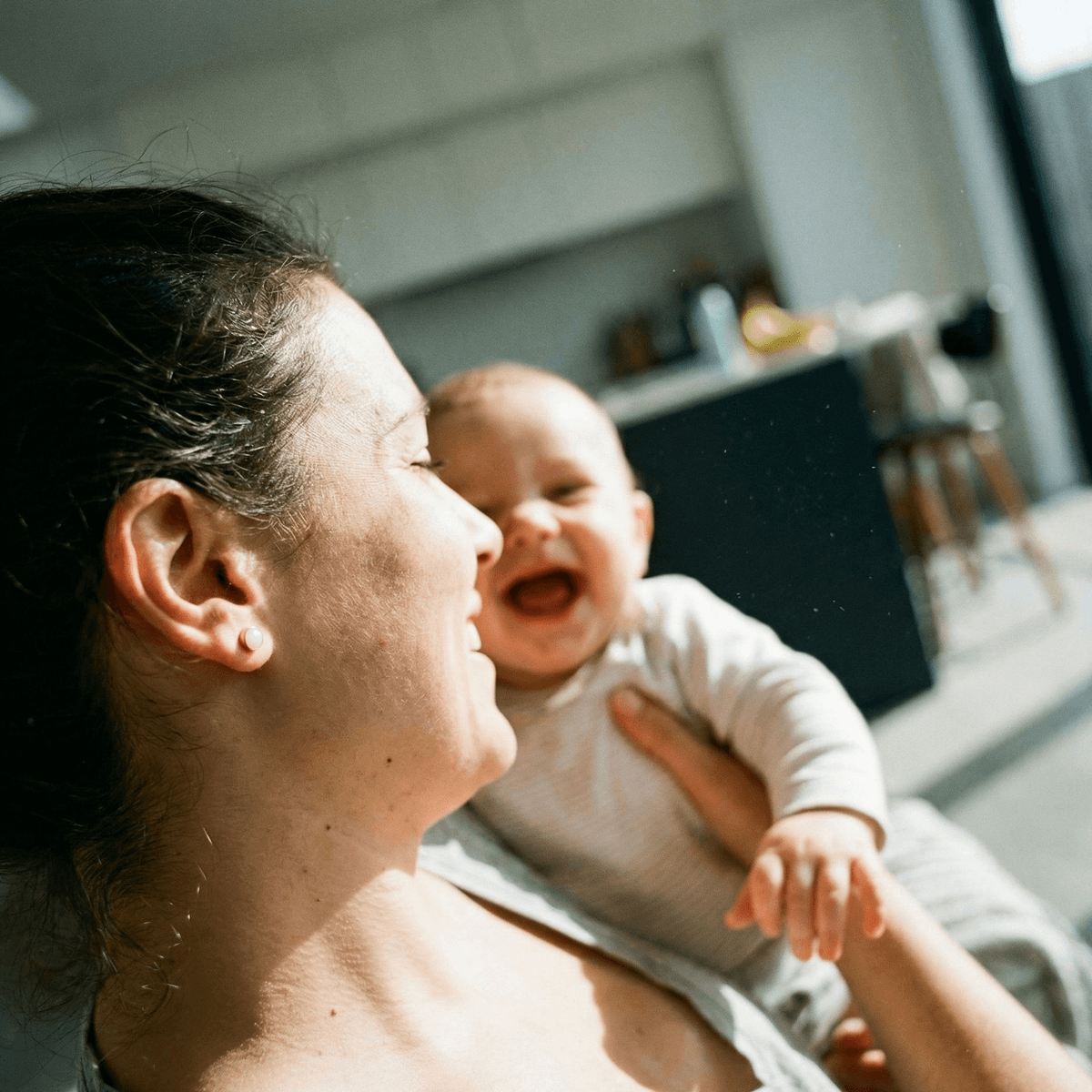 Sunlit home scene of a mother cuddling and laughing with her baby, wearing small stud earrings that resemble a diy breastmilk earring kit keepsake.