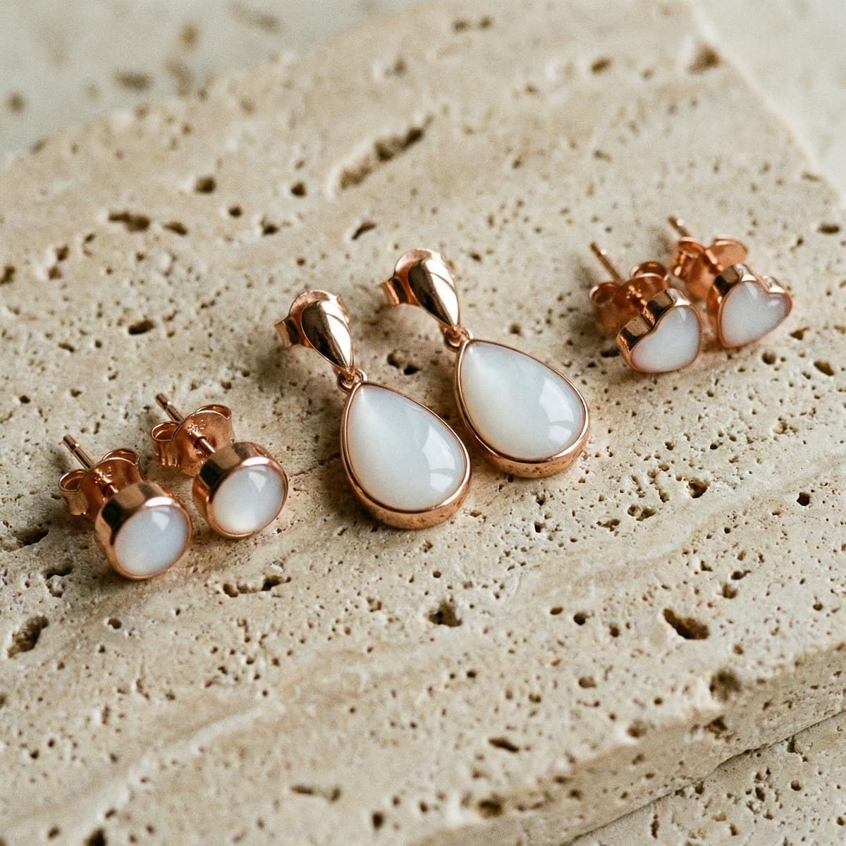 Rose gold jewelry set on a textured beige stone background, featuring milky white resin teardrop and heart-shaped diy breastmilk earrings with matching pieces.
