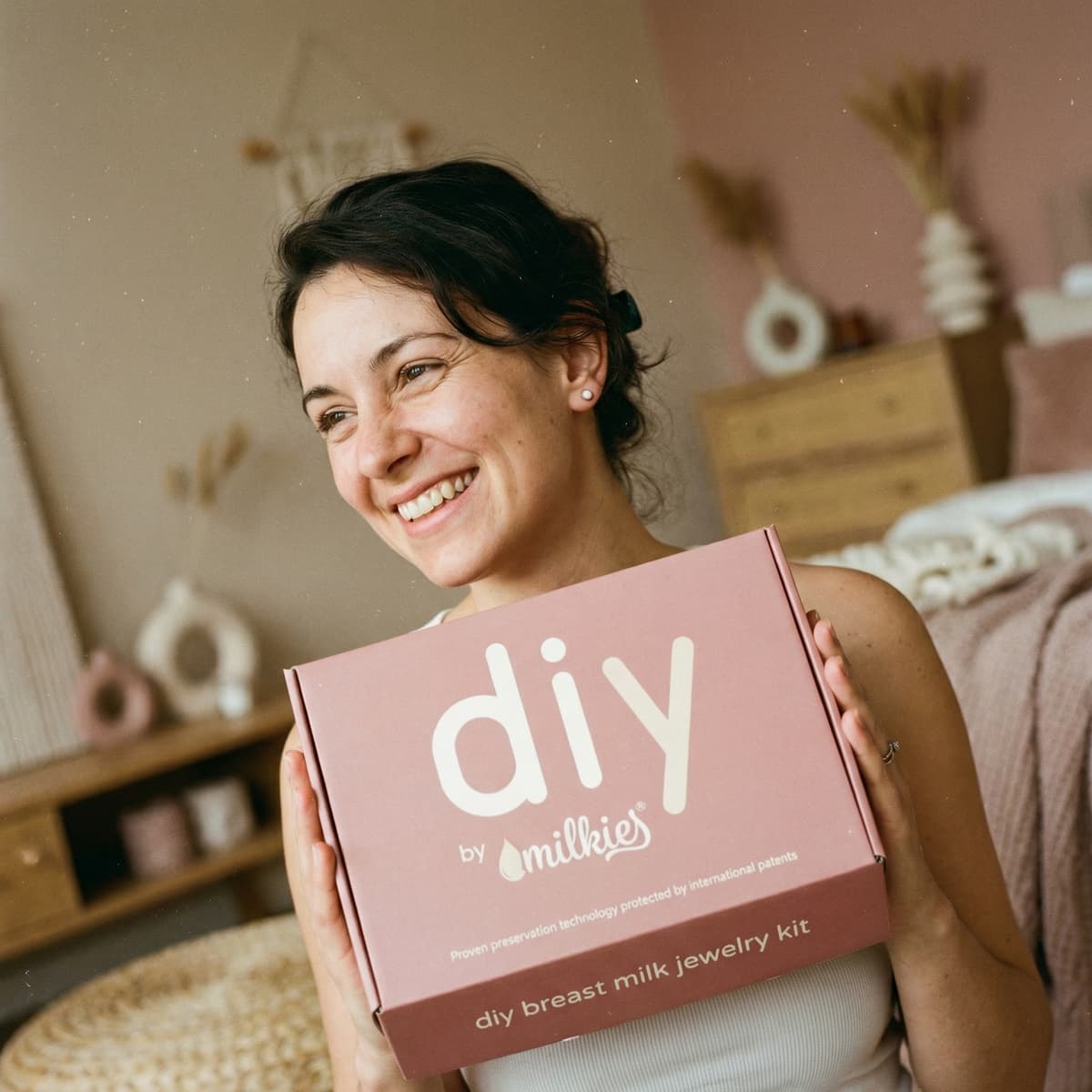 Smiling mom holding a pink DIY by MILKIES kit box for making diy breastmilk earrings at home, highlighting patented preservation technology and why an at-home keepsake kit is a smart choice.