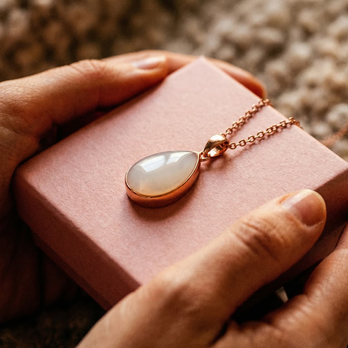 Hands holding a pink gift box with a rose-gold teardrop keepsake pendant, showing why a diy breastmilk necklace kit is a meaningful at-home way to create a personal memento.