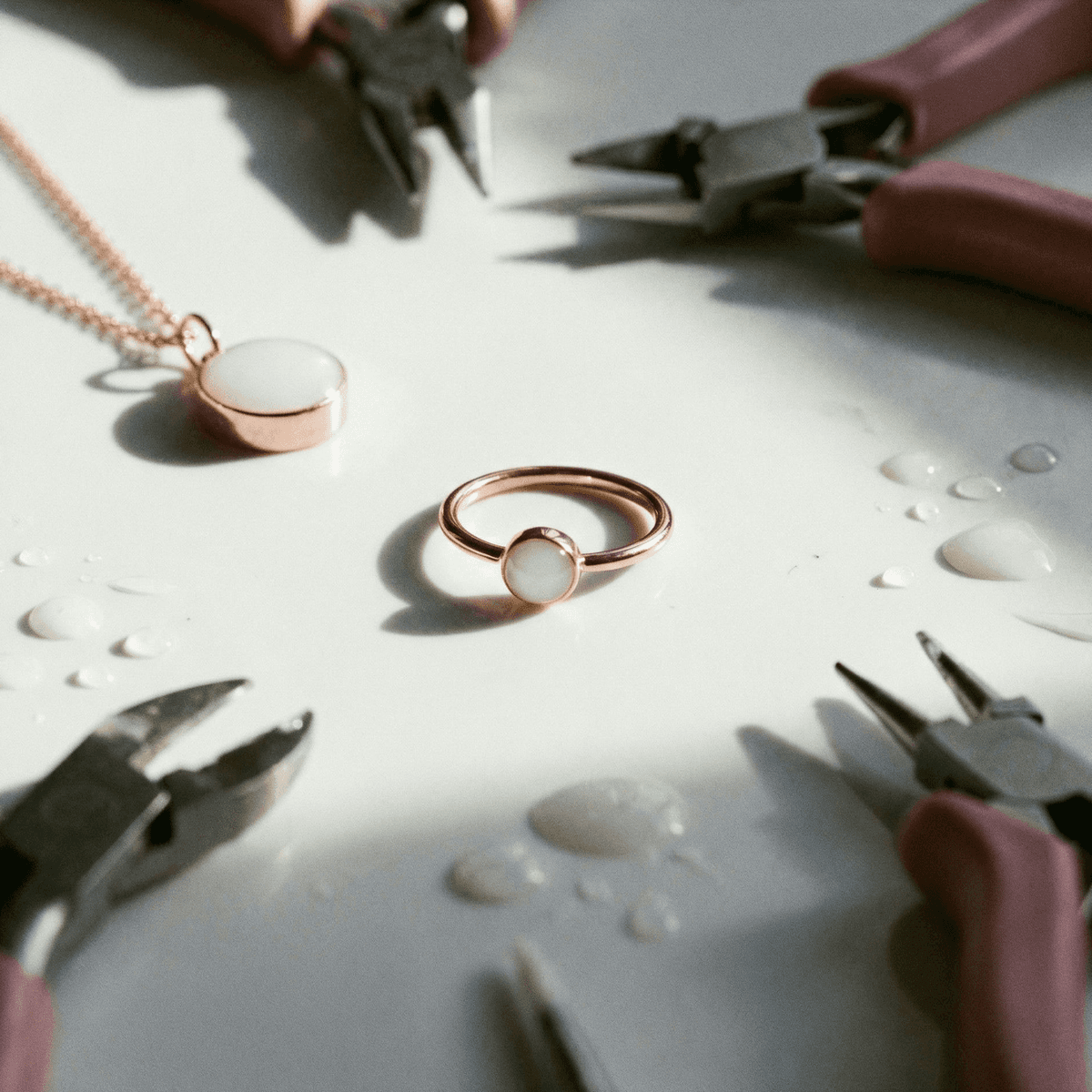 Hands using jewelry pliers to assemble a rose-gold diy breastmilk ring on a white work surface, with a matching pendant necklace and small drops of milk nearby.