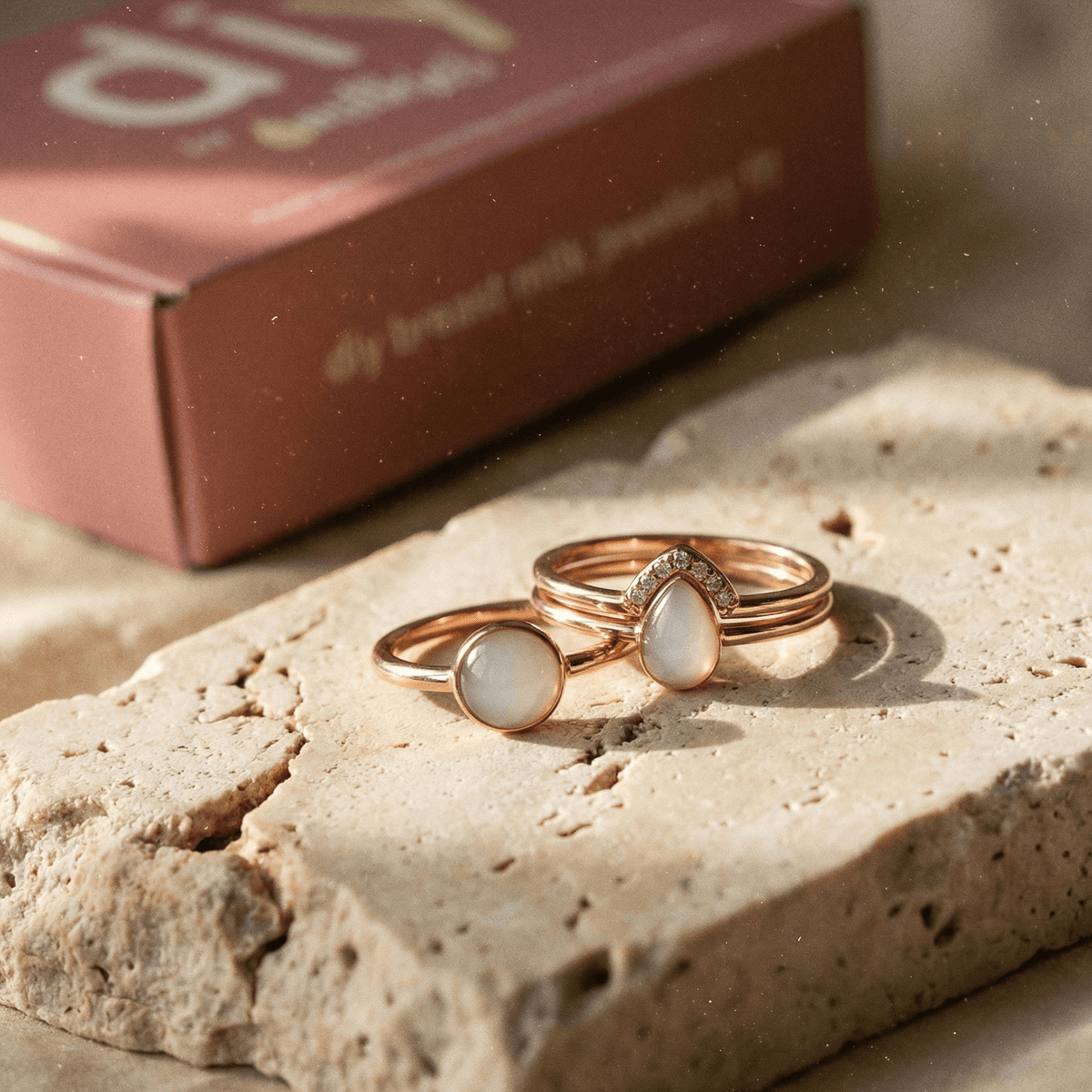 Rose gold rings with milky-white stones displayed on textured stone, featuring a DIY by MILKIES diy breastmilk ring kit keepsake jewelry set with a blurred kit box in the background.