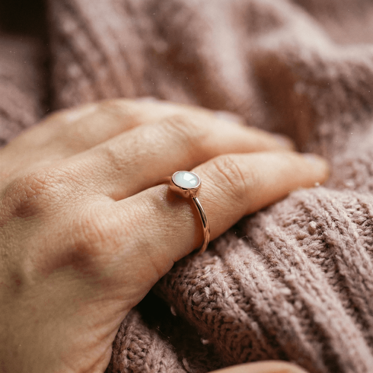 Close-up of a hand wearing a rose-gold ring with a milky white resin stone, showcasing diy breastmilk rings on a soft pink knitted blanket.